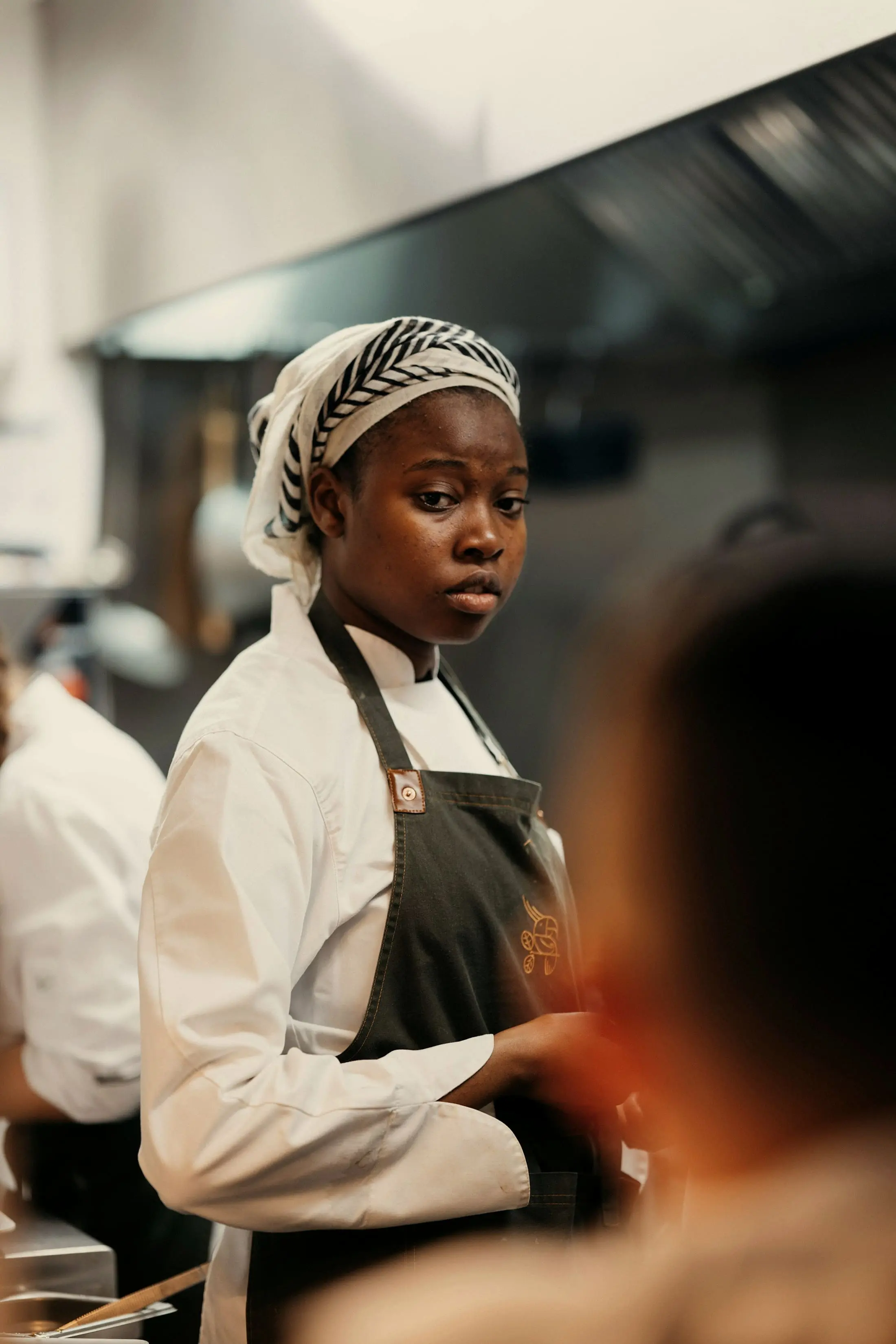 A cook in a white jacket and dark apron, head wrapped, looking calmly to camera