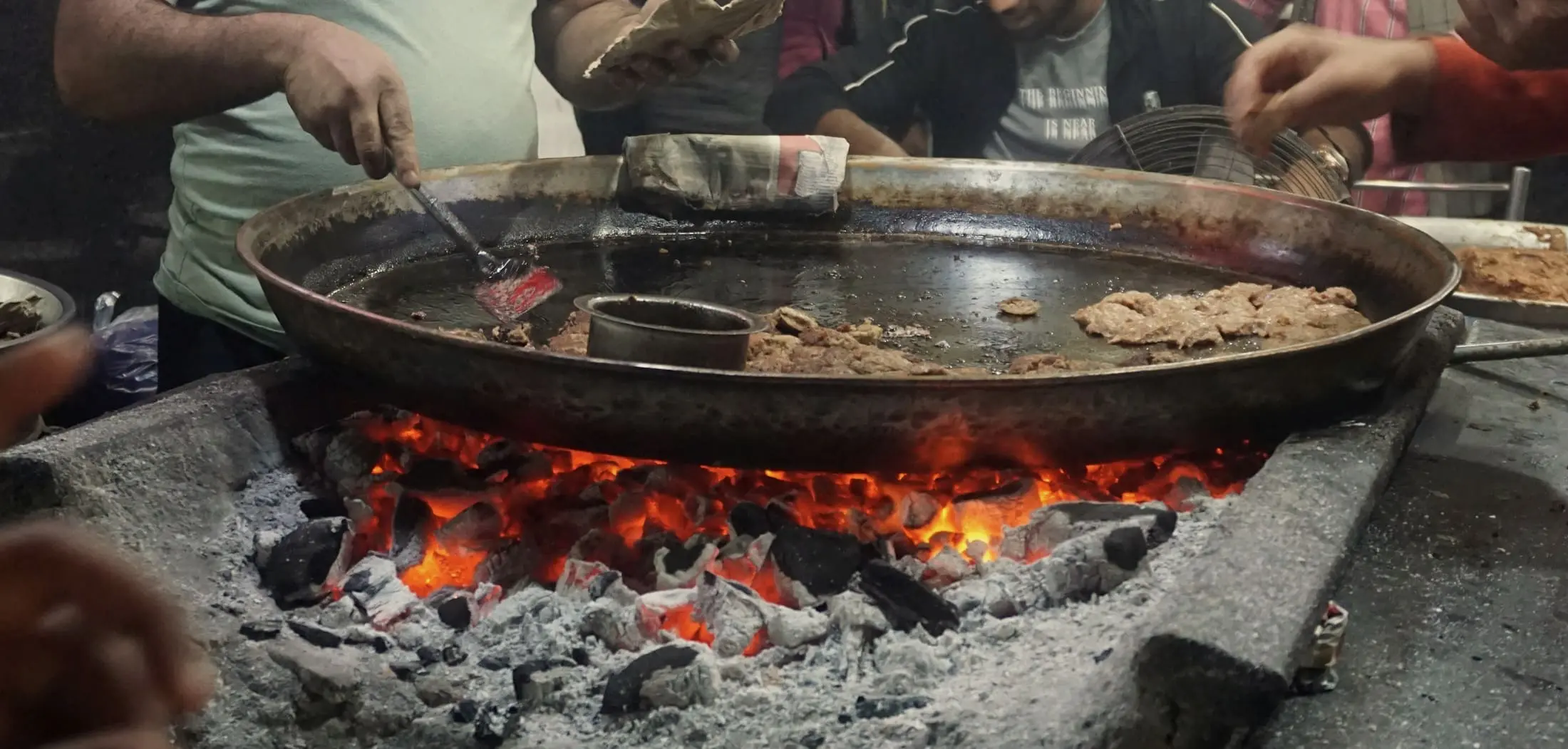 A wide steel pan resting above glowing charcoal and grey ash