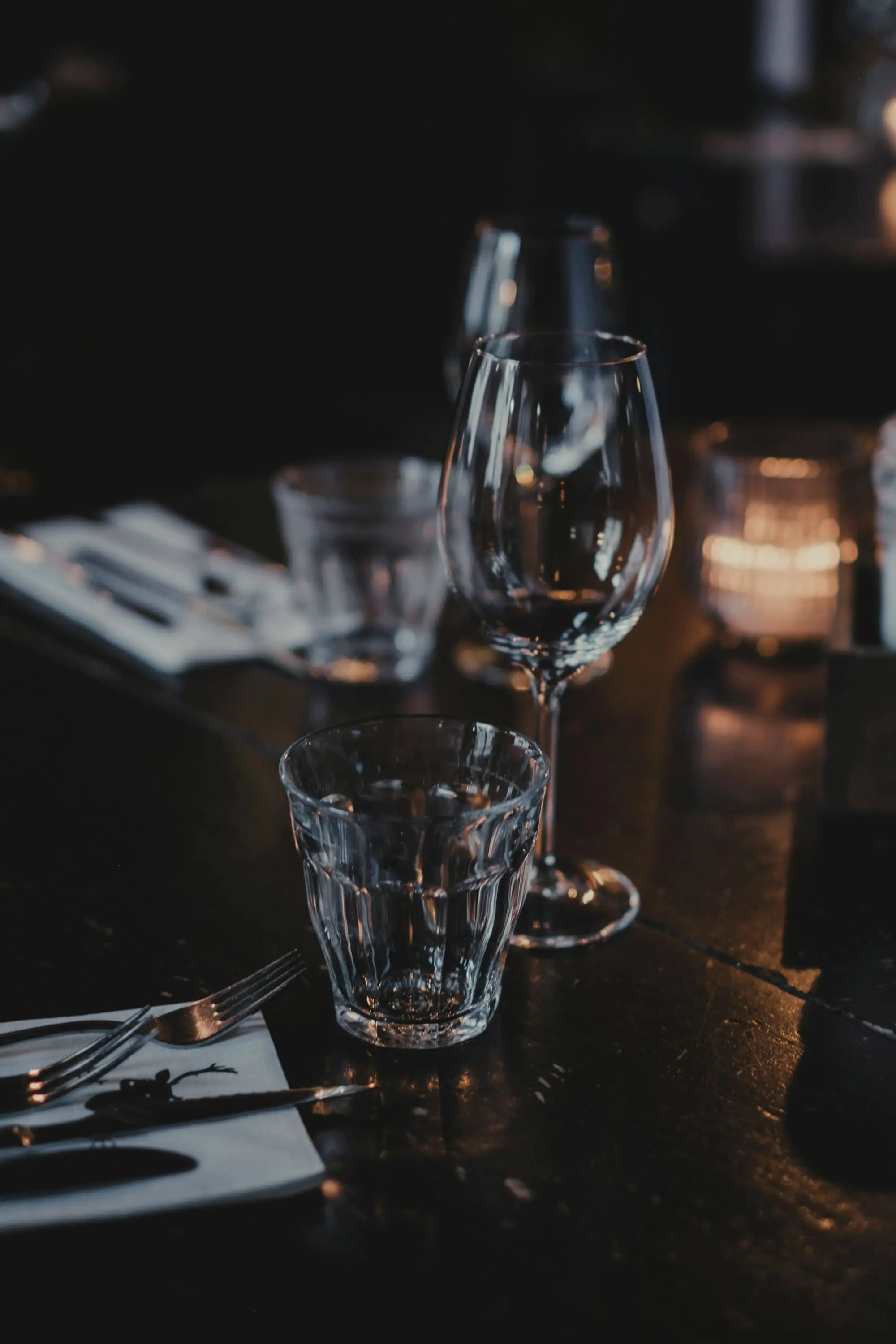 Wine glasses and a tumbler on a table, a candle burning behind