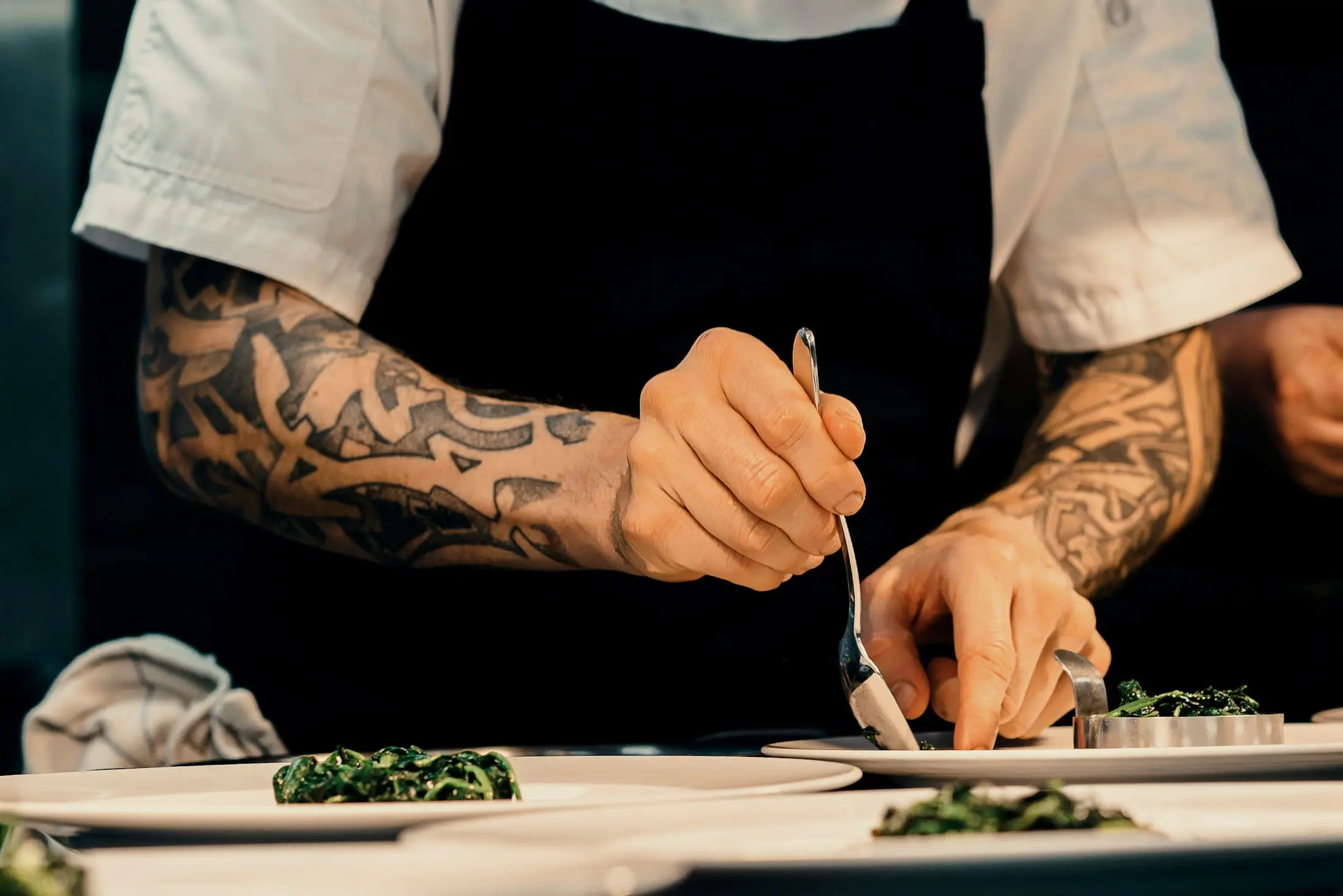 Tattooed hands placing a garnish with tweezers on a plate